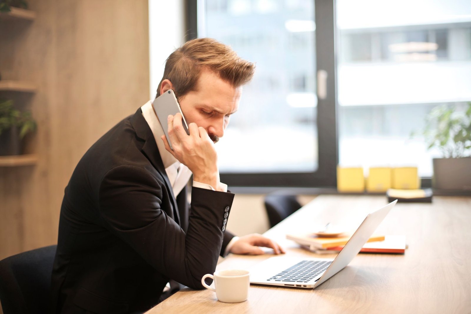 A man on a phone call at his desk while checking something on his laptop