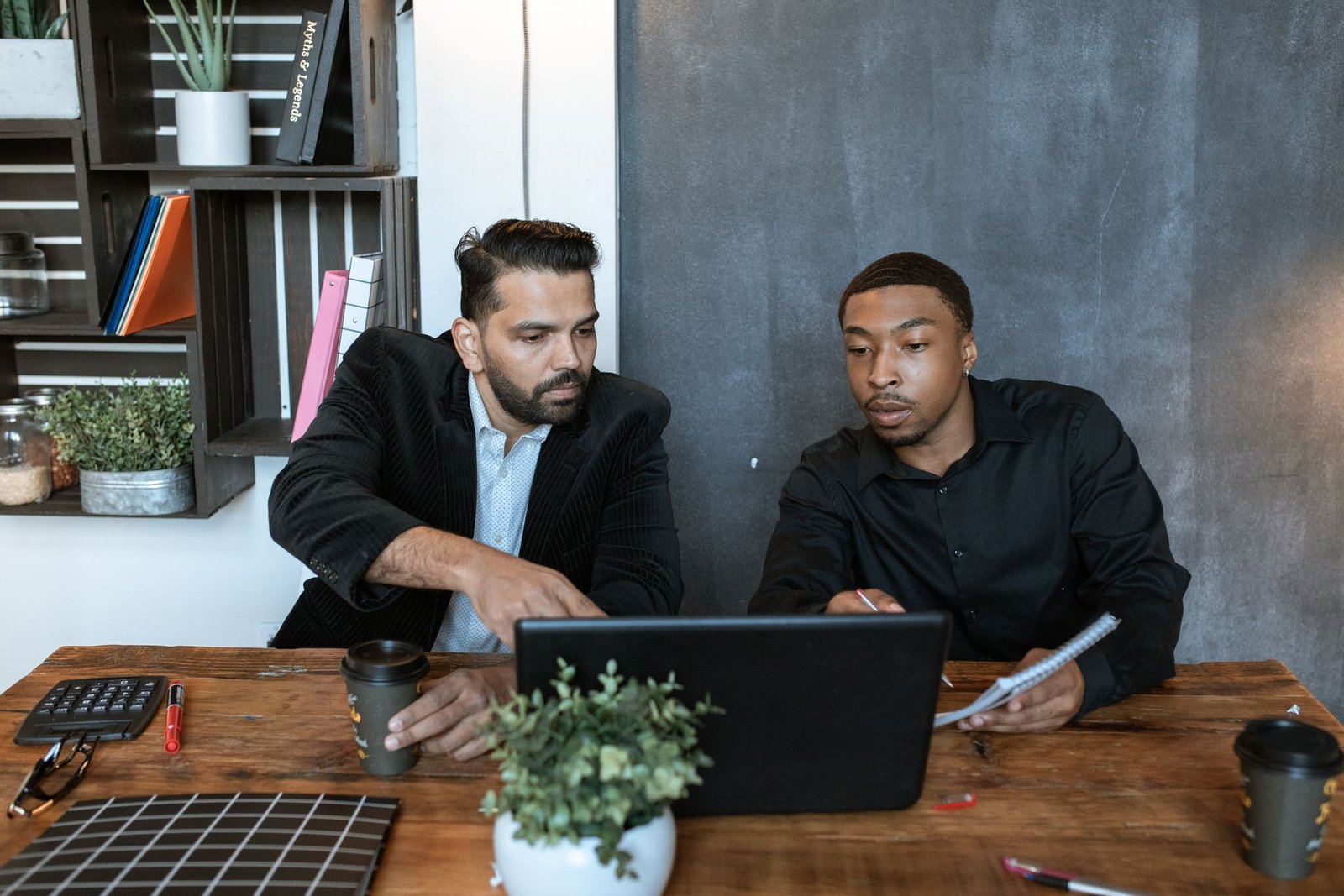 Two colleagues at a wooden desk reviewing work together on a laptop